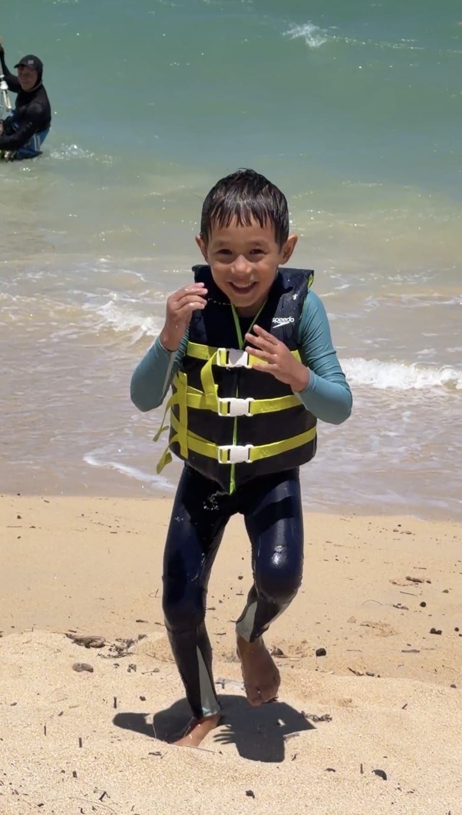 Aiden walking out of the water in his life jacket, smiling, with Andrey landing the kite in the shallows behind him